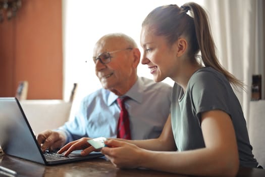 pexels-photo-3823488-3823488 Young woman in casual clothes helping senior man in formal shirt with paying credit card in Internet using laptop while sitting at table
